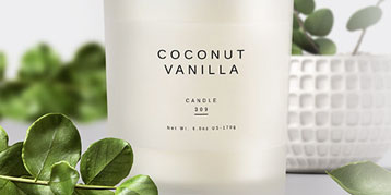 Minimalist white candle with a white square label on a table with pot plant in the background