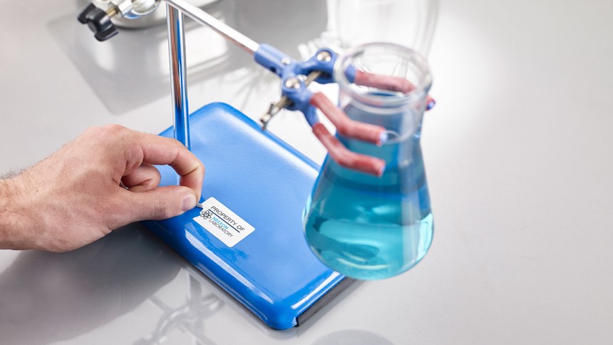 Person's hands trying to peel off labels off a blue surface that are not coming off properly. There is a beaker with blue liquid in the foreground.