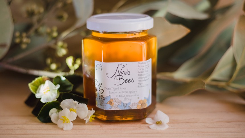 Honey jar with printed label on the front. Sitting on a wooden surface next to small flowers and green leaves.