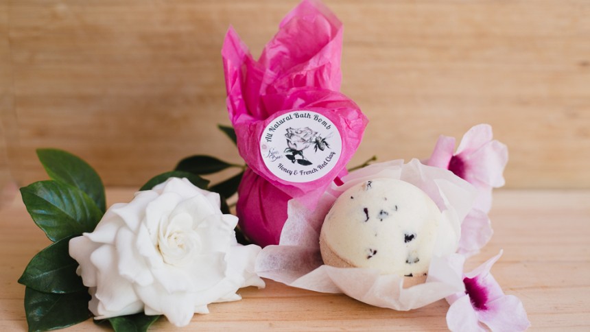 A cream ball shaped material sitting on white tissue paper in front of a wrapped item in pink tissue paper with a white round label. Sitting next to a white flower with green leaves.