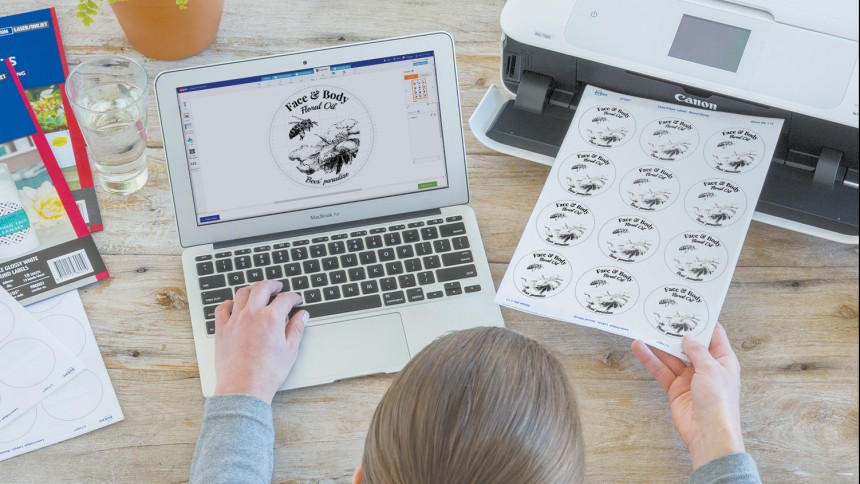 A person in front of a laptop which using their left hand to type. The right hand holds a sheet of labels that looks as if it has just been pulled out of a printer.