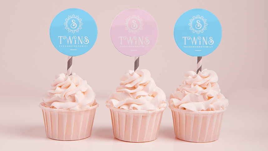 Three cupcakes with light pink frosting in pink wrappers. Each cupcake has a striped straw with a circular topper. The topper on the left is blue, the one in the middle is pink, and the one on the right is blue. All the toppers say "TWINS" with the words "THE SANDOZ FAMILY" below and a stylized letter S above. The background is a pale pink color.