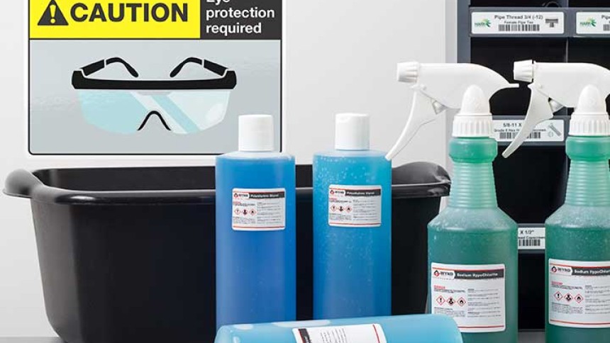 A tabletop scene with several bottles of blue and green cleaning solutions, a black bin, spray bottles, and a caution sign that reads "CAUTION Eye protection required" with an icon of safety goggles. In the background, there is a storage unit with labeled drawers.