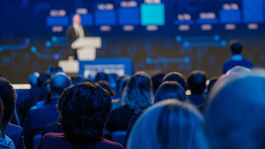 A crowd of people, seen from behind, are seated in an auditorium listening to a speaker on a stage. The speaker is a man in a suit, standing behind a podium. The large screen behind him is blurry, but shows a blue graph. The room is dimly lit with blue light.
