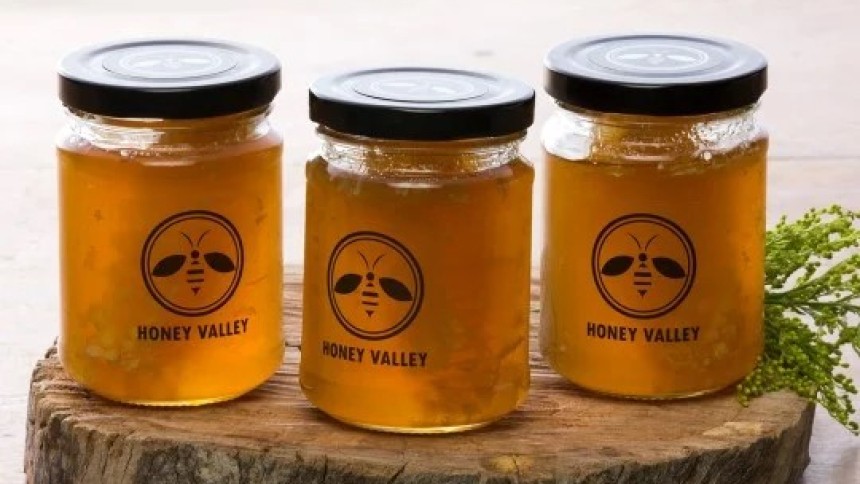 Three glass jars filled with golden honey, each labeled with a round logo featuring a bee and the words “Pure Honey.” They are placed on a wooden slab with a sprig of greenery beside them.