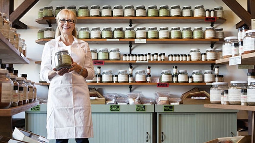 Woman working in a health food store holding a large glass jar with dried herbs or spice with a green label on the jar container