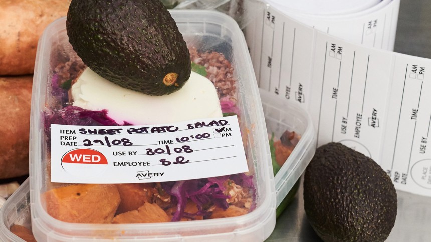Food rotation labels with day of the week and fields to label items for fresh food with food rotation labels in a roll and salad and fresh vegetables in the background.