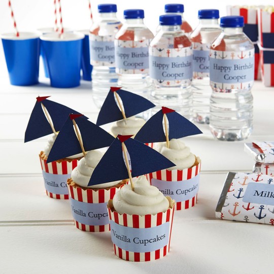 A party table with a nautical theme is shown, displaying various snacks and drinks. There are six cupcakes in red and white striped wrappers, each with a blue sail topper. Two "Milk Choc Bars" are wrapped in anchor-patterned paper. In the background, there are water bottles with custom "Happy Birthday Cooper" labels, blue paper cups with red striped straws, and popcorn boxes.
