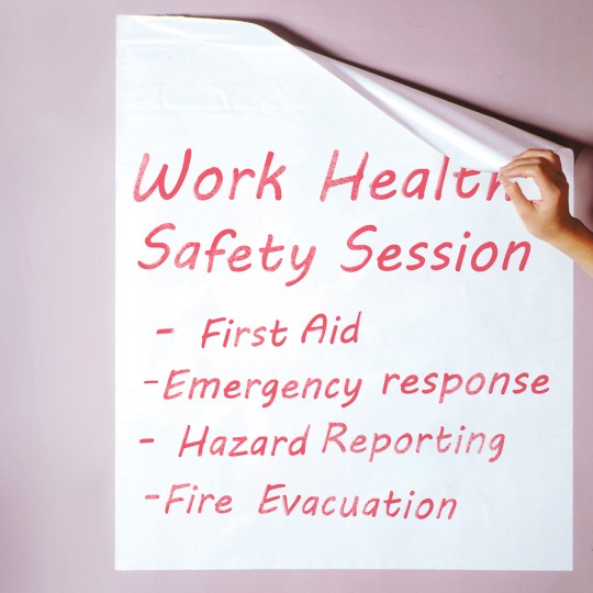 A white sheet of paper with red text is being peeled off a wall. The text reads "Work Health Safety Session" followed by a list of four bullet points: "First Aid," "Emergency response," "Hazard Reporting," and "Fire Evacuation."