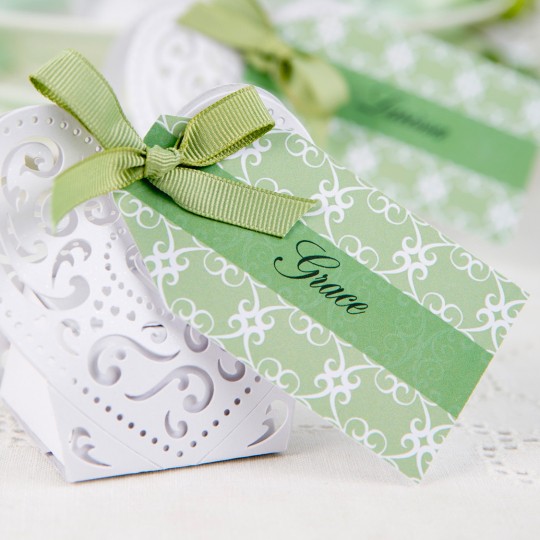 A small, white, laser-cut heart-shaped box is shown on a white lace tablecloth. A green ribbon is tied in a bow on top of the box. A green and white tag with a swirling pattern is attached to the ribbon, and the name "Grace" is written in black script on the tag.
