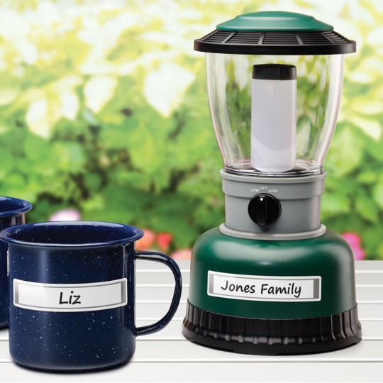 A green and black camping lantern with a clear globe and a white cylindrical light is positioned on a white wooden table. Two dark blue speckled camping mugs sit next to it. All three items have white labels on them: the lantern is labeled "Jones Family," and the mugs are labeled "Mark" and "Liz." The background is a blurry green garden.