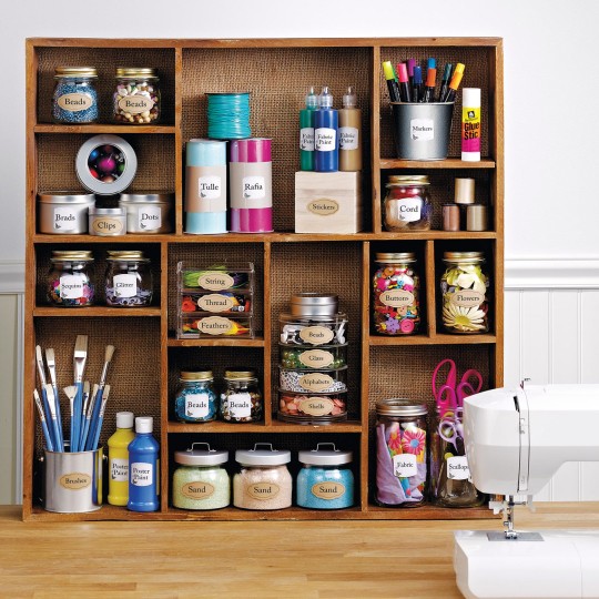 A rustic wooden shelving unit with a burlap back is filled with various craft supplies in jars, containers, and bottles. On the shelves are items like beads, clips, glitter, threads, sand, and fabric swatches, all neatly labeled. A sewing machine, paint brushes, paint bottles, markers, and scissors are also visible on the wooden table in front of the unit.