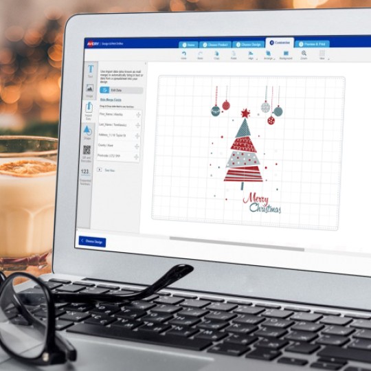 Laptop sitting on desk with christmas decorations around it. On screen is Avery Design and Print platform with a Christmas label design on display.