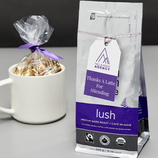 Conference table setting. Gift of a mug with chocolates and a bag of coffe on the table. On the coffee bag is a personalised printed tag with "Thanks a latte for attending"