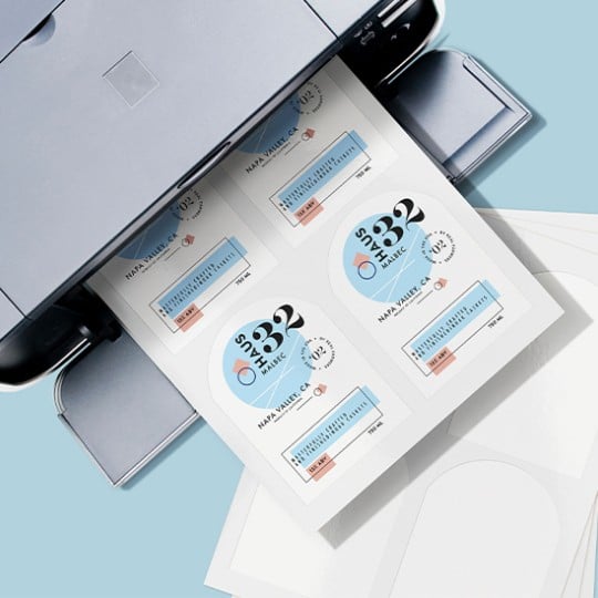 A close-up, high-angle shot of a printer ejecting a sheet of paper with multiple wine bottle labels printed on it. The labels are predominantly white and light blue with black text, featuring "HAUS" as a prominent brand name, "MALBEC," "NAPA VALLEY, CA," and other wine details. Partially visible blank label sheets are stacked below.