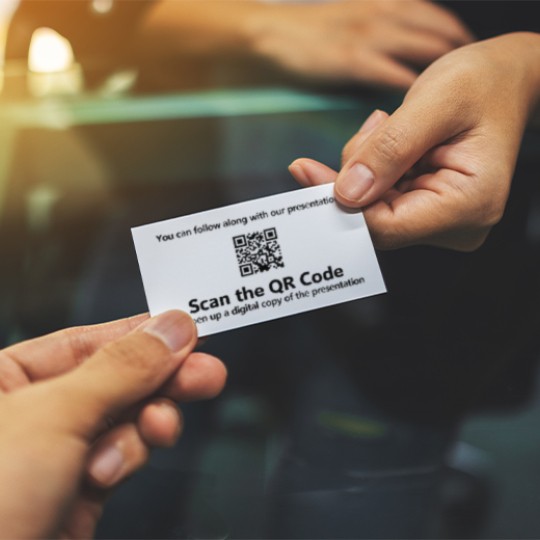 Two hands are shown, one handing a white card with a QR code and the text "Scan the QR Code" to another, implying the exchange of information for a presentation.
