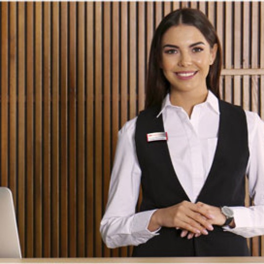 A smiling women with long dark hair, in a white shirt and black vest wearing a name tag/ She is standing in front of a wooden wall with vertical slats.