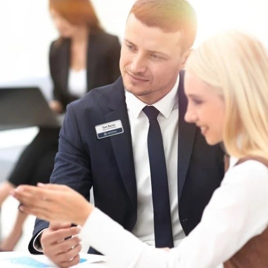 A man in a suit with a name badge assisting a women with a white shirt and long blond hair.