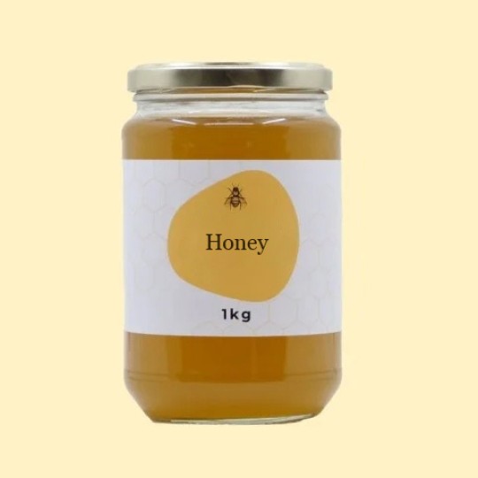 A front view of a large, transparent glass jar of honey with a silver lid. The label is white with a repeating honeycomb pattern. In the center of the label, there is an abstract, golden-yellow shape with a small illustration of a bee and the text "Honey". Below this, the text "1kg" is printed. The jar is set against a plain, light yellow background.