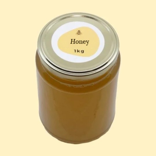 A high-angle view of a large, transparent glass jar of honey with a silver lid. A circular white label is centered on the top of the lid, featuring an abstract, golden-yellow shape with a small illustration of a bee and the text "Honey". Below this, the text "1kg" is printed. The jar is set against a plain, light yellow background.