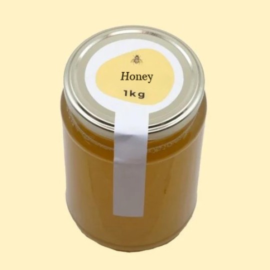 A high-angle view of a large, transparent glass jar of honey with a silver lid. A white security sticker is placed over the lid and extends down the side of the jar. A circular white label is centered on the top of the lid, featuring an abstract, golden-yellow shape with a small illustration of a bee and the text "Honey". Below this, the text "1kg" is printed. The jar is set against a plain, light yellow background.