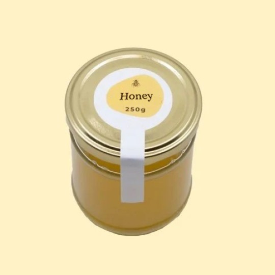 A high-angle view of a short, wide, transparent glass jar of honey with a gold lid. A white security sticker is placed over the lid and extends down the side of the jar. A circular white label is centered on the top of the lid, featuring an abstract, golden-yellow shape with a small illustration of a bee and the text "Honey". Below this, the text "250g" is printed. The jar is set against a plain, light yellow background.
