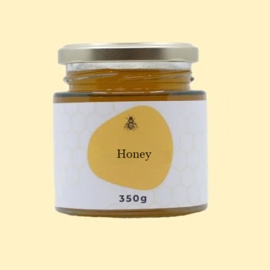 A front view of a short, wide, transparent glass jar of honey with a silver lid. The label is white with a repeating honeycomb pattern. In the center of the label, there is an abstract, golden-yellow shape with a small illustration of a bee and the text "Honey". Below this, the text "350g" is printed. The jar is set against a plain, light yellow background.