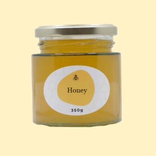 A front view of a short, wide, transparent glass jar of honey with a silver lid. The label is a white oval with a repeating honeycomb pattern. In the center of the label, there is an abstract, golden-yellow shape with a small illustration of a bee and the text "Honey". Below this, the text "350g" is printed. The jar is set against a plain, light yellow background.