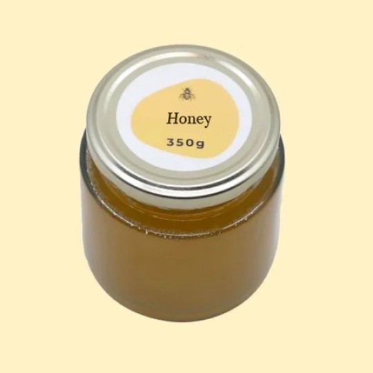 A high-angle view of a short, wide, transparent glass jar of honey with a silver lid. A circular white label is centered on the top of the lid, featuring an abstract, golden-yellow shape with a small illustration of a bee and the text "Honey". Below this, the text "350g" is printed. The jar is set against a plain, light yellow background.