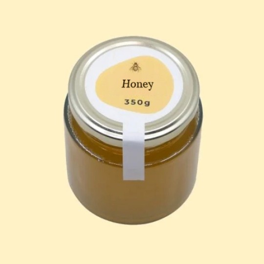 A high-angle view of a short, wide, transparent glass jar of honey with a silver lid. A white security sticker is placed over the lid and extends down the side of the jar. A circular white label is centered on the top of the lid, featuring an abstract, golden-yellow shape with a small illustration of a bee and the text "Honey". Below this, the text "350g" is printed. The jar is set against a plain, light yellow background.