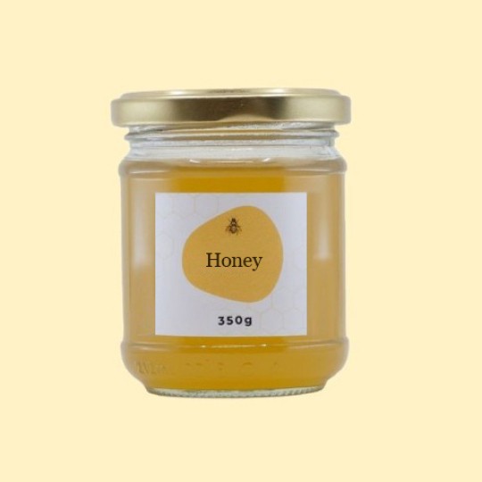 A front view of a short, wide, transparent glass jar of honey with a gold lid. The label is white with a repeating honeycomb pattern. In the center of the label, there is an abstract, golden-yellow shape with a small illustration of a bee and the text "Honey". Below this, the text "350g" is printed. The jar is set against a plain, light yellow background.