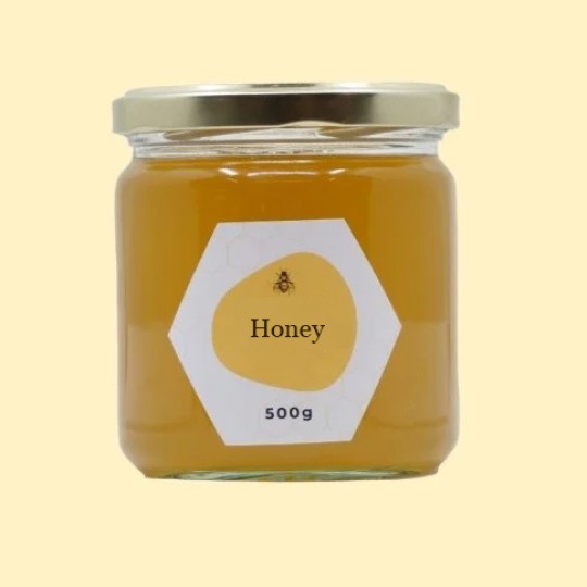A front view of a short, wide, transparent glass jar of honey with a gold lid. The label is a white hexagon. In the center of the label, there is an abstract, golden-yellow shape with a small illustration of a bee and the text "Honey". Below this, the text "500g" is printed. The jar is set against a plain, light yellow background.