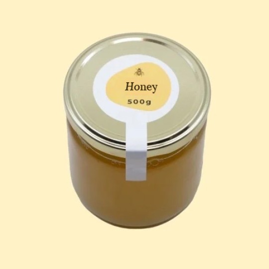 A high-angle view of a large, transparent glass jar of honey with a gold lid. A white security sticker is placed over the lid and extends down the side of the jar. A circular white label is centered on the top of the lid, featuring an abstract, golden-yellow shape with a small illustration of a bee and the text "Honey". Below this, the text "500g" is printed. The jar is set against a plain, light yellow background.