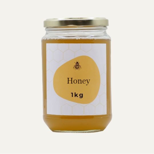 A large, transparent glass jar of honey with a gold lid. The label is white with a repeating honeycomb pattern. In the center of the label, there is an abstract, golden-yellow shape with a small illustration of a bee and the text "Honey". Below this, the text "1kg" is printed. The jar is set against a plain, light background.
