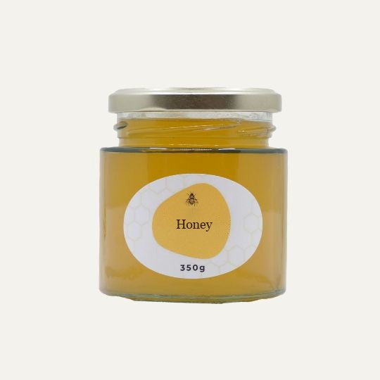 A wide, transparent glass jar of honey with a silver lid. The label is a white oval with a repeating honeycomb pattern. In the center of the label, there is an abstract, golden-yellow shape with a small illustration of a bee and the text "Honey". Below this, the text "350g" is printed. The jar is set against a plain, light background.