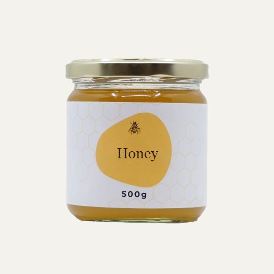A small, transparent glass jar of honey with a gold lid. The label is white with a repeating honeycomb pattern. In the center of the label, there is an abstract, golden-yellow shape with a small illustration of a bee and the text "Honey". Below this, the text "500g" is printed. The jar is set against a plain, light background.