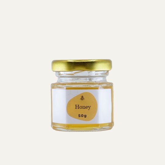 A miniature, transparent glass jar of honey with a gold lid. The label is white with a repeating honeycomb pattern. In the center of the label, there is an abstract, golden-yellow shape with a small illustration of a bee and the text "Honey". Below this, the text "50g" is printed. The jar is set against a plain, light background.