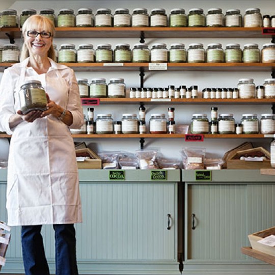 Woman working in a health food store holding a large glass jar with dried herbs or spice with a green label on the jar container
