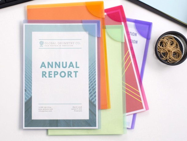 A top-down view of a white desk with a keyboard, a laptop, and a small cup of rubber bands. In the center, a stack of colorful semi-transparent plastic folders holds a document titled "ANNUAL REPORT" for "GLOBAL GEOMETRY CO." The folders are orange, pink, light green, and purple. The "Annual Report" cover page has a blue and green geometric design.