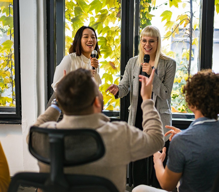 This image features a group of employees singing with microphones. They appear to be partaking in karaoke and are in a well lit room with greenery outside of the window. There are two women singing and two men in the audience cheering