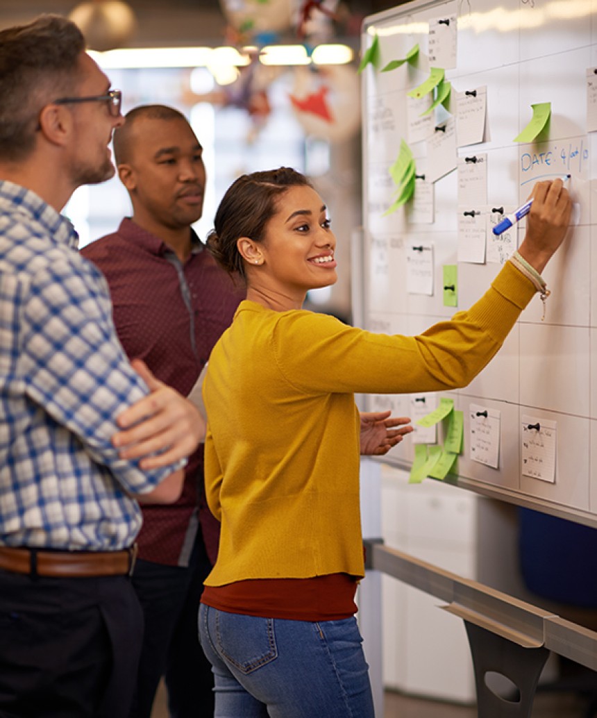 This image features three people in a corporate office setting. There is a woman in the front smiling and writing on a white board. The white board is covered with sever notes and other important information, the team appear to be working together on some sort of planner for their company
