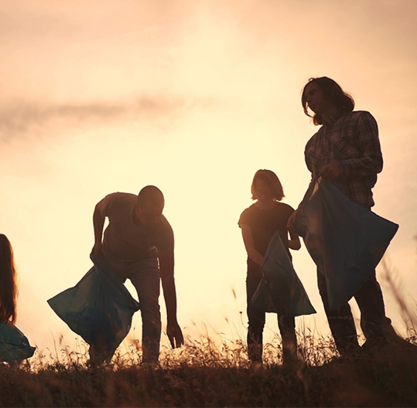This image features a group of people out in a field holding plastic rubbish bags and picking up rubbish. It suggests they are a team and cleaning up the field as a team bonding activity.