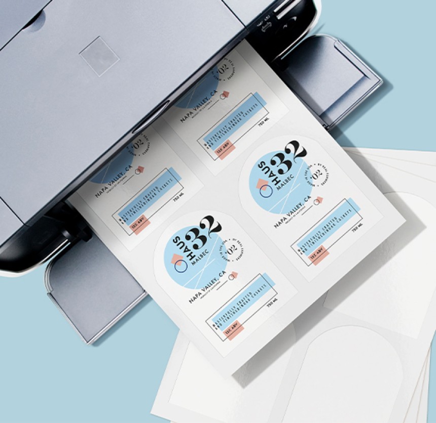 A close-up, high-angle shot of a printer ejecting a sheet of paper with multiple wine bottle labels printed on it. The labels are predominantly white and light blue with black text, featuring "HAUS" as a prominent brand name, "MALBEC," "NAPA VALLEY, CA," and other wine details. Partially visible blank label sheets are stacked below.