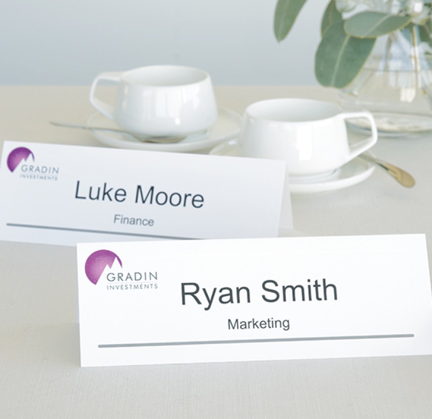 This image features two name cards on a conference table. The names on the card read "Ryan Smith" from Marketing and "Luke Moore" from finance. Both cards feature a company logo with a pink and white mountain and the company is called Gradin Investments. Behind these cards are two sets of tea cups and plates