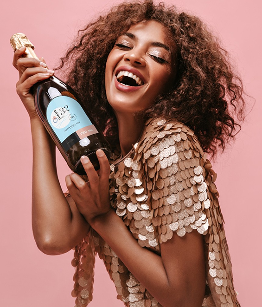A smiling woman with voluminous curly brown hair and a sparkling gold sequined top holds a bottle of sparkling wine, looking joyful against a plain pink background.