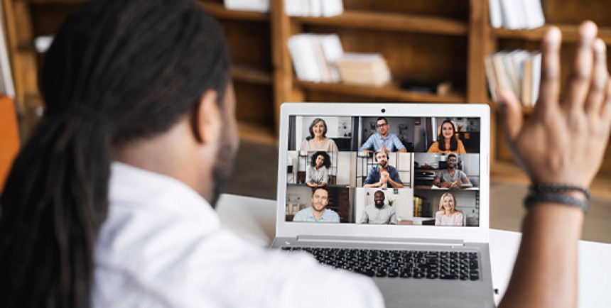 A man with long dreadlocks is seen from behind, sitting at a desk and waving at his laptop screen during a video call. The laptop shows a grid of diverse people participating in a virtual meeting. In the background, there are bookshelves and a warm, indoor setting, suggesting a home or library environment.