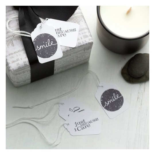 The image features a beautifully wrapped gift with a black ribbon, accompanied by several gift tags. The tags display messages like 'smile' and 'just because I care' along with decorative elements. A lit candle and smooth stones are also visible in the composition, enhancing a serene atmosphere.