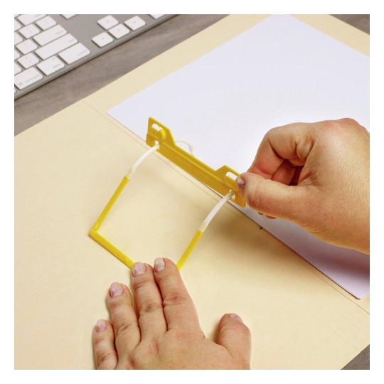 The image shows a person's hand using a yellow tool or clip to hold a piece of white paper against a tan background, likely for some kind of organizational or crafting purpose. A keyboard is visible in the background, suggesting this is a workspace.