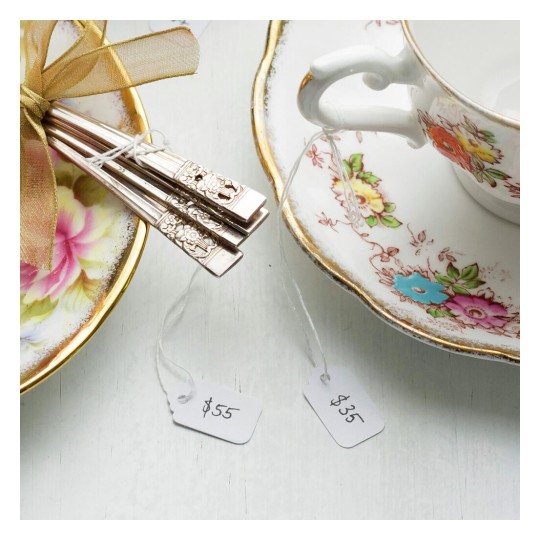 The image features a decorative plate with silver-colored utensils tied with a bow, and a delicate teacup beside it. Price tags indicate $55 and $35.