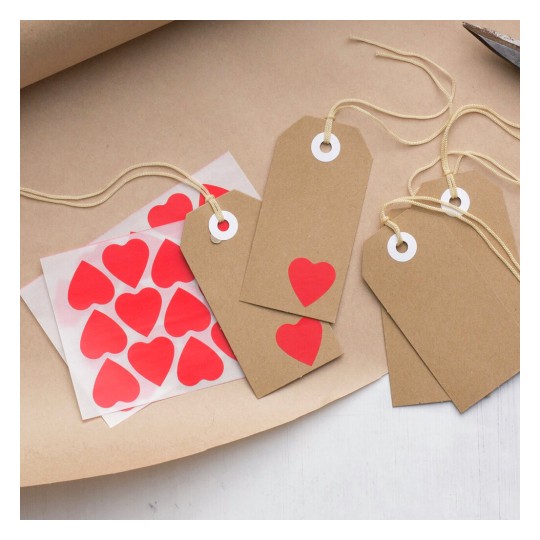 The image features brown gift tags with red heart stickers, some tags are blank while one has a heart cutout. There is also a sheet of red heart-shaped stickers in the background on craft paper.